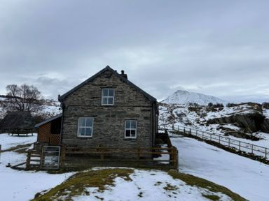 A stone house with snow around and mountains in the background at Ty Siabod in Capel Curig near Betws-Y-Coed