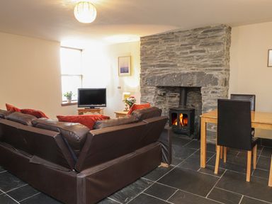 A living room with a brown leather sofa red cushions stone fireplace wood stove television and wooden dining table with black chairs at Ty Llewelyn in Capel Curig near Betws Y Coed