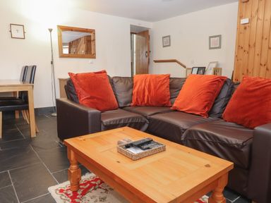 A living room with a brown leather sofa and orange cushions a wooden coffee table a wooden dining table with black chairs a floor lamp a wall mirror and wooden door at Ty Llewelyn in Capel Curig near Betws-Y-Coed