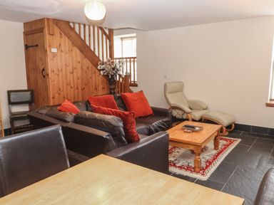 A sitting area with a black leather sofa with red cushions a wooden coffee table on a rug and a beige armchair with an ottoman near wooden stairs at Ty Llewelyn in Capel Curig near Betws-Y-Coed