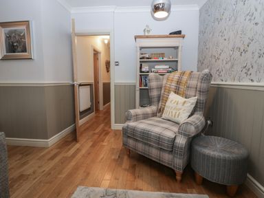 A plaid armchair with a pillow and throw blanket next to a wicker ottoman and bookshelf in a room at 1 Boarbank Farm in Allithwaite near Cartmel