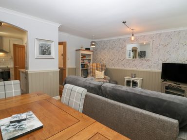 A living room with a wooden dining table and plaid chairs a gray sofa a patterned wingback chair a bookshelf a TV and a mirror at 1 Boarbank Farm in Allithwaite near Cartmel