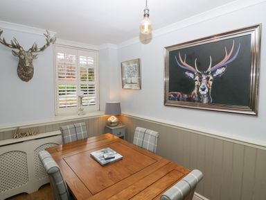 A dining room with a wooden table and four plaid chairs with a deer head wall mount and a large deer painting at 1 Boarbank Farm in Allithwaite near Cartmel