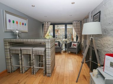 A room with a wicker bar table and stools two armchairs with decorated cushions a tripod floor lamp wooden floor and glass doors at 1 Boarbank Farm in Allithwaite near Cartmel