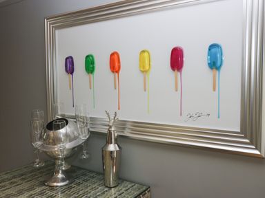 A framed artwork of colorful popsicles melting on a wall above a table with a silver ice bucket holding glasses and a silver cocktail shaker at 1 Boarbank Farm in Allithwaite near Cartmel