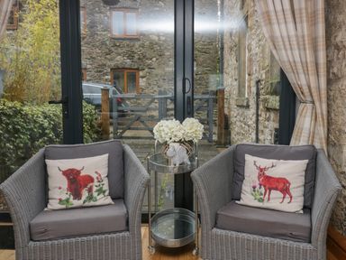 Two wicker armchairs with cushions and decorative animal pillows by glass doors and a small round table with flowers at 1 Boarbank Farm in Allithwaite near Cartmel