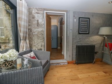 A room with a wicker chair with pillows, a glass window with plaid curtains, a floor lamp, a wall radiator, and a doorway showing a refrigerator at 1 Boarbank Farm Allithwaite near Cartmel