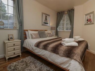 A bedroom with a bed covered in a brown blanket and decorative pillows nightstands with lamps and framed pictures and windows with curtains at 1 Boarbank Farm in Allithwaite near Cartmel