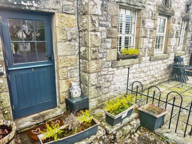An outdoor stone patio area with a blue door numbered 1 flower pots with plants a small black table with a snowman decoration and a black metal fence at 1 Boarbank Farm in Allithwaite near Cartmel