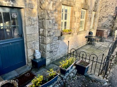 A stone building exterior with a blue door a small black table with a snowman figure flowerpots with plants a fenced stone patio with a round glass table and stacked black chairs at 1 Boarbank Farm Allithwaite near Cartmel