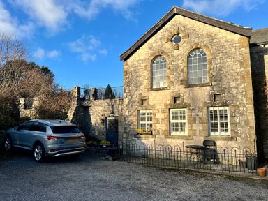 An exterior view of a stone building with arched windows a parked silver car and a small fenced patio with a table and chair at 1 Boarbank Farm Allithwaite near Cartmel