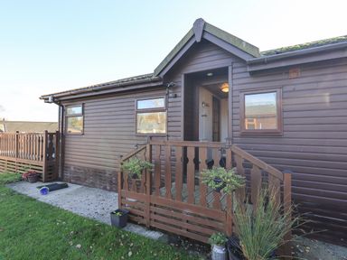 The exterior of a single-story wooden house with two windows and a small fenced porch with hanging plants at Kellet Lodge Lakeside 26 Dock Acres near Carnforth