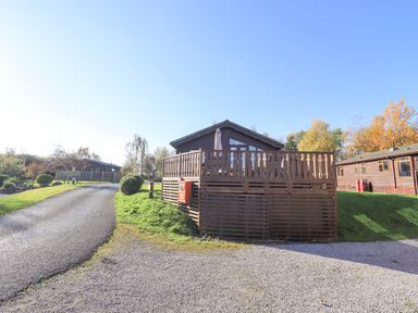 A wooden lodge with a fenced porch next to a paved road and grassy area at Kellet Lodge Lakeside 26 in Dock Acres near Carnforth