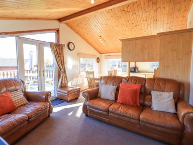 A living room with leather sofas and cushions near glass doors leading to a deck with outdoor furniture at Kellet Lodge Lakeside 26 Dock Acres near Carnforth