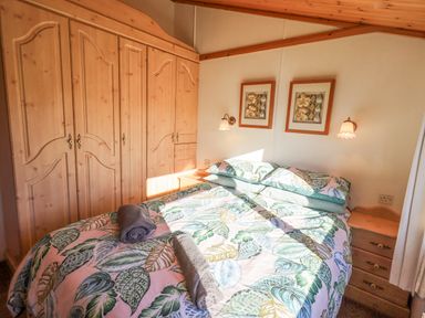 A bedroom with a double bed with leaf patterned bedding two rolled towels on top wooden wardrobes two framed pictures and bedside tables at Kellet Lodge Lakeside 26 Dock Acres near Carnforth