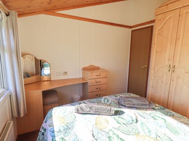 A bedroom with a bed covered in leaf patterned bedding towels on the bed a wooden wardrobe a dressing table with mirror and stool at Kellet Lodge Lakeside 26 Dock Acres near Carnforth