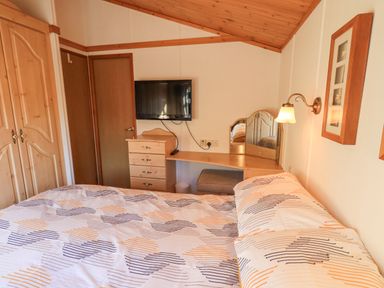 A bedroom with a bed a wall-mounted tv a wooden dresser a vanity desk with a mirror and a wall lamp at Kellet Lodge Lakeside 26 Dock Acres near Carnforth