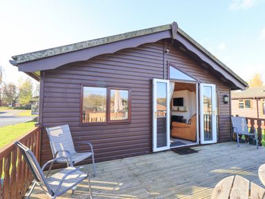 A deck with chairs outside a wooden cabin with open double doors showing a living room at Kellet Lodge Lakeside 26 Dock Acres near Carnforth