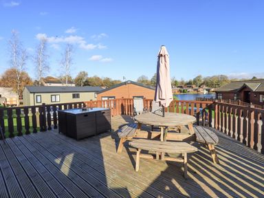 An outdoor wooden deck with a round picnic table and umbrella surrounded by wooden railings and overlooking cabins and a lake at Kellet Lodge Lakeside 26 in Dock Acres near Carnforth