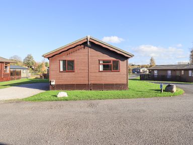 A brown wooden cabin with two windows on a grassy plot near a paved road at Kellet Lodge Lakeside 26 Dock Acres near Carnforth