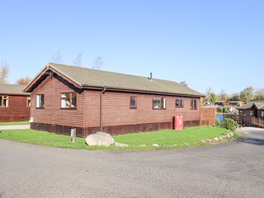 A wooden cabin with multiple windows on a grassy area near a paved road at Kellet Lodge Lakeside 26 Dock Acres near Carnforth