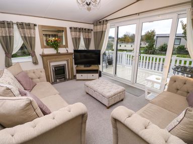 A living room with two sofas a patterned ottoman a fireplace a TV on a stand and large sliding glass doors at 117 Crosswinds Hillway near Bembridge