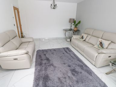 A living room with two beige sofas a round side table with a lamp and plant and a gray rug on white tiled floor at Golygfa Hafan in Little Haven