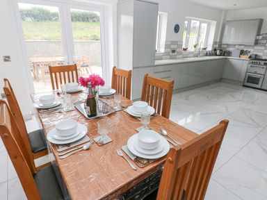 A dining table set for six with white dishes and glasses in a kitchen with grey cabinets and white tiled floor at Golygfa Hafan in Little Haven