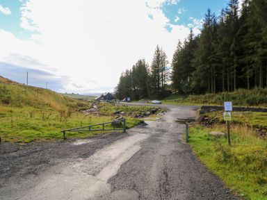 A rural road leading into a forested area with parked cars and a small building at The Museum in Nenthead