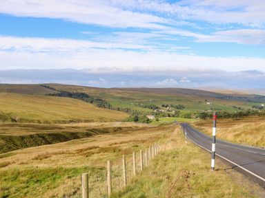 A rural road running alongside a fence and grassy fields with hills in the distance at The Museum in Nenthead