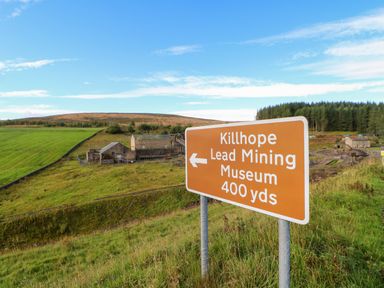 A sign pointing to Killhope Lead Mining Museum with buildings and grassy hills in the background at The Museum in Nenthead