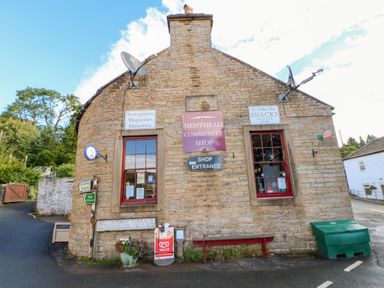 A stone building with signs for Nenthead Community Shop newspapers magazines groceries and snacks with two windows and a bench outside at The Museum in Nenthead