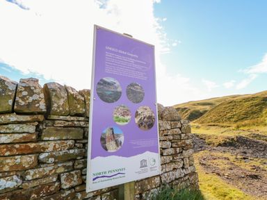 A stone wall with a purple informational sign about UNESCO Global Geoparks in a grassy hilly outdoor area at The Museum in Nenthead