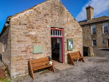 A stone building with two wooden benches outside and an open door showing interior at The Museum in Nenthead