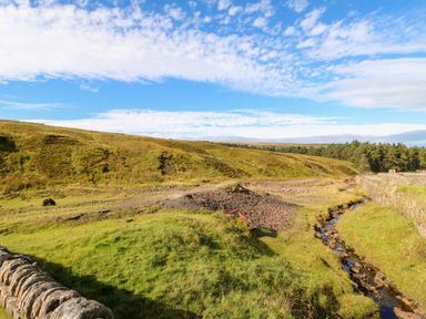 A grassy hillside with a small stream running alongside a stone wall at The Museum in Nenthead