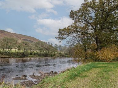 A river flowing beside a grassy bank with trees and hills in the background at Bradan Lodge in Dalmally