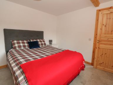A bedroom with a double bed with plaid bedding a red blanket a dark gray headboard a wooden door and a small white bedside table with a lamp at Bradan Lodge in Dalmally