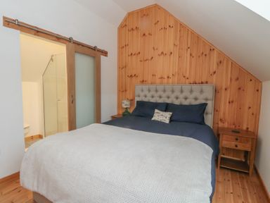 A bedroom with a bed with a gray headboard and navy bedding next to a wooden nightstand and a sliding wooden door leading to a bathroom at Bradan Lodge in Dalmally