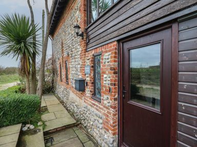 The exterior of a stone and brick house with a wooden door and windows and a pathway with plants at Bridge Barn in Sidestrand near Overstrand