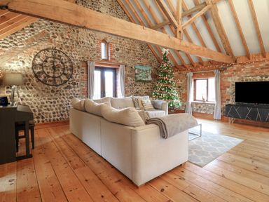 A living room with a beige sofa a Christmas tree a large wall clock a piano a television and wooden beams at Bridge Barn in Sidestrand near Overstrand