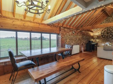 An open room with wooden beams a dining table bench chairs a window overlooking a field and a seating area with a piano at Bridge Barn in Sidestrand near Overstrand