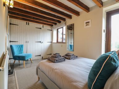 A bedroom with a bed, folded towels, a blue chair, wooden ceiling beams, a window, and a mirror at Bridge Barn in Sidestrand near Overstrand