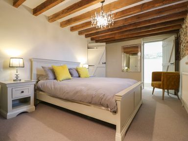 A bedroom with a bed with gray bedding and yellow pillows two bedside tables with lamps a mustard armchair a mirror wooden ceiling beams and a door at Bridge Barn in Sidestrand near Overstrand