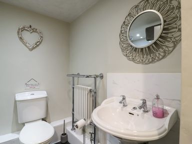 A bathroom with a toilet a pedestal sink with two taps a round decorative mirror and a heart-shaped wall decoration at Bridge Barn in Sidestrand near Overstrand