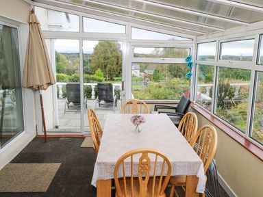 A dining area with a table and six wooden chairs in a glass sunroom overlooking a garden at Lakeview Lodge in Knockmore County Mayo