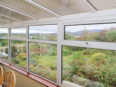 A glass-enclosed sunroom with wooden chairs overlooking a garden and distant mountains at Lakeview Lodge in Knockmore County Mayo