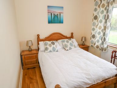 A bedroom with a wooden bed frame white bedding two leaf patterned cushions two wooden bedside tables each with a lamp and a window with leaf patterned curtains at Lakeview Lodge in Knockmore County Mayo