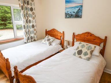 A bedroom with two wooden single beds white bedding leaf patterned pillows and curtains a window and a seaside painting on the wall at Lakeview Lodge in Knockmore County Mayo