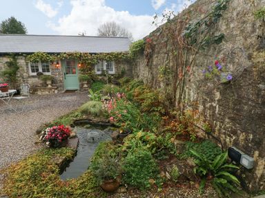 A garden with a small pond and flowers next to a stone wall and a stone building with a green door and windows at Little Cloud in Nash near Cowbridge