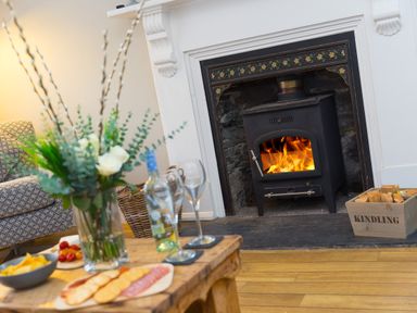 A living room with a wood burning stove with fire a wooden table with snacks wine bottle and glasses and a vase with flowers at Captains Walk in Newquay
